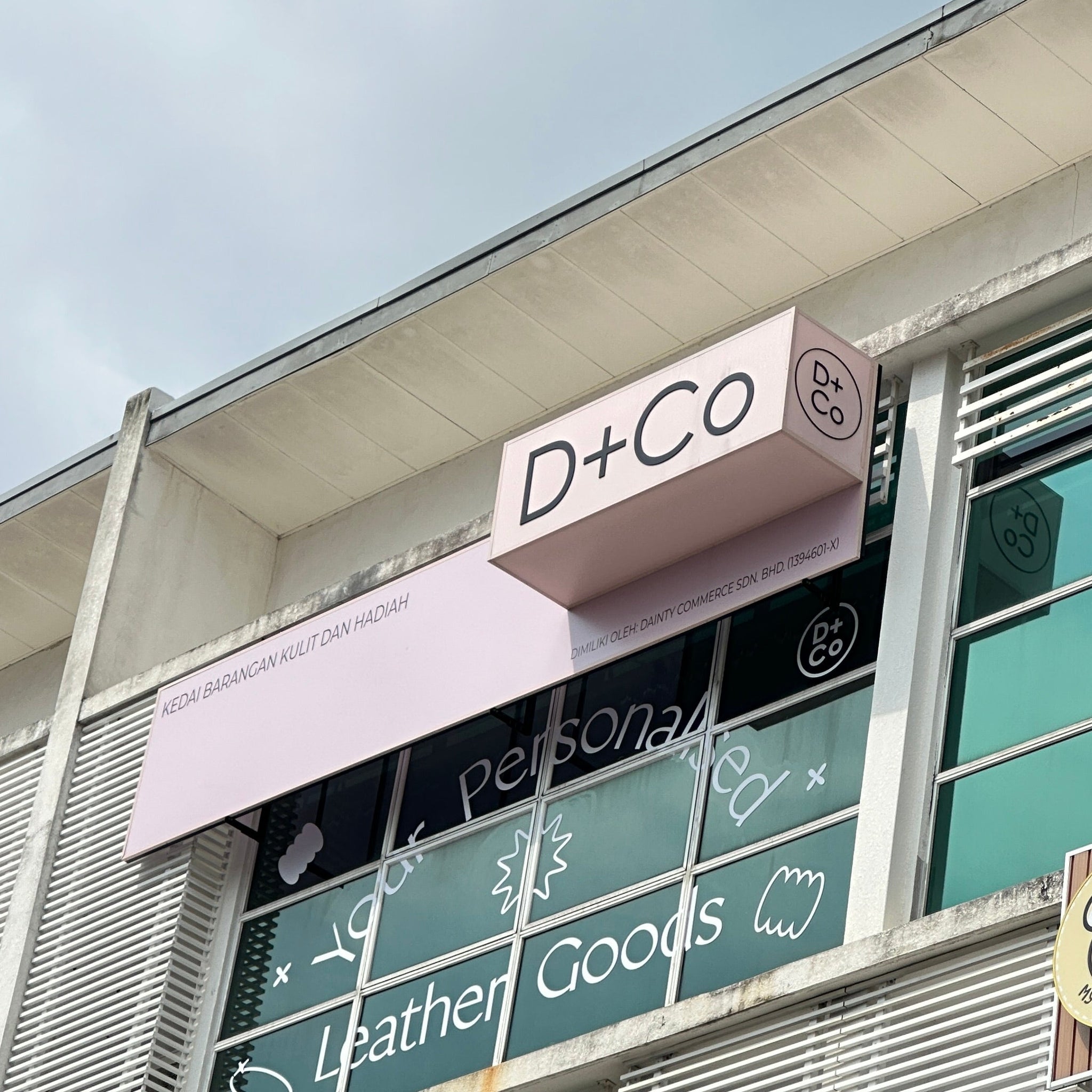 A low-angle shot of a building facade featuring a pink 'D+Co' sign above glass windows with 'Personalised Leather Goods' written on them.