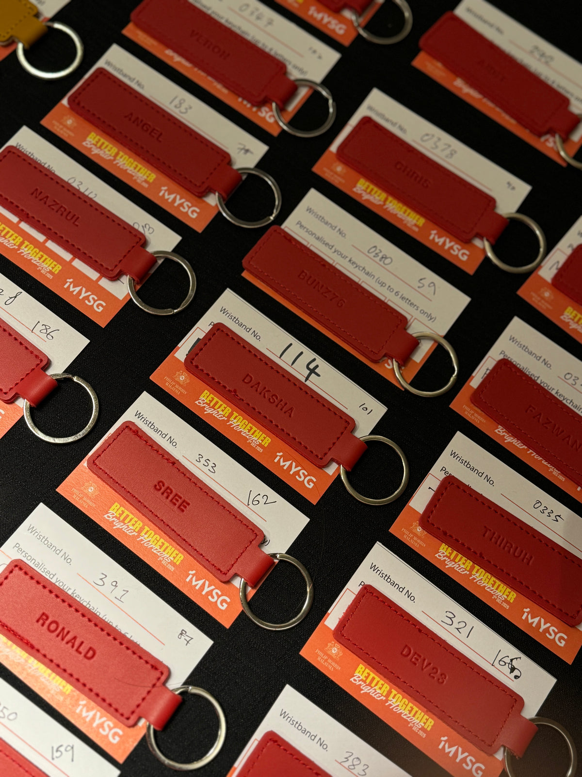 Multiple red leather keychains with silver metal key rings, each attached to a white and orange card printed with passenger-style details and the text MYSG, arranged diagonally on a dark background.